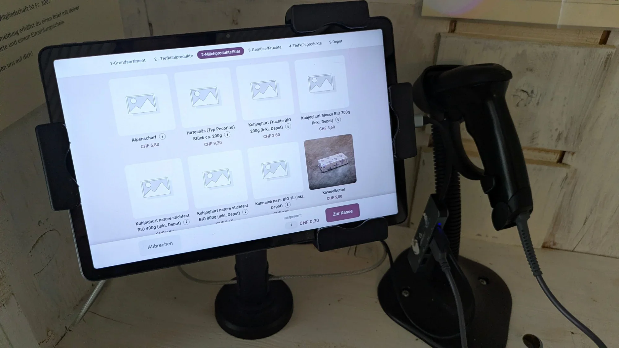 Tablet mit Scanner am Kassenplatz, Bildschirm zeigt Milchprodukte und Eier mit Preisen, darunter Alpenscharf und Hirtech&auml;s