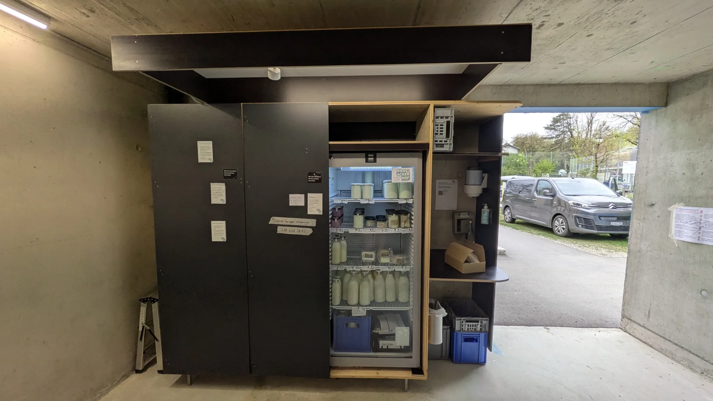 Schwarzer Schrank mit K&uuml;hlschrank, Regalen und Milchharassen im Vorraum der Velogarage