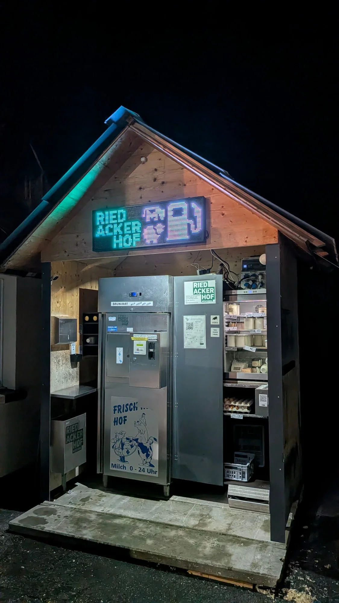 Beleuchtetes Holzh&auml;uschen mit Milchautomat, K&uuml;hlschrank mit Joghurt, K&auml;se und Eiern bei Nacht